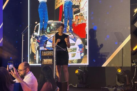 Penny Mallory giving a motivational speech on stage at the NCE Awards. There is a projection of a racing car behind her.