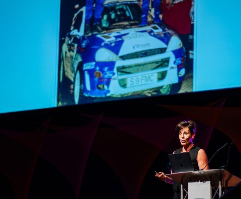 Female motivational speaker Penny Mallory on stage with picture of world rally car behind her on the screen