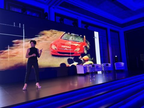 Image of Penny Mallory stood onstage giving a motivational speech with blue lighting. There is an image of a race car projected behind her.