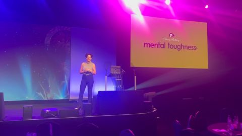 Image of Penny Mallory presenting her Mental Toughness slideshow on stage with blue and pink surrounding lighting.