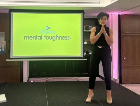 Image of Penny Mallory stood on stage giving a motivational speech with a mental toughness presentation in the background.